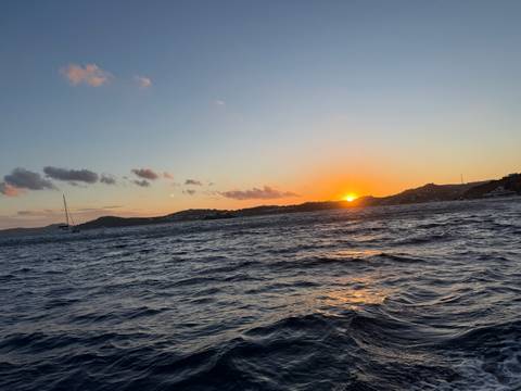       Sun setting over choppy sea waters with distant hills and a lone sailboat
  