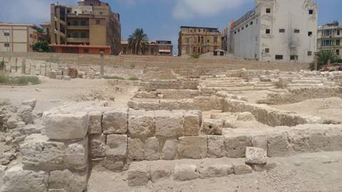       Stone foundations of an archaeological excavation site with city buildings in the background under a blue midday sky.
  