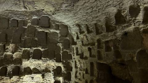       Dimly lit interior of an ancient rock-cut chamber with rows of carved niches in the walls.
  