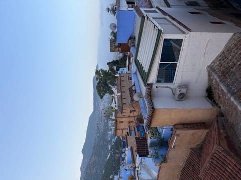       Rooftop view over blue and earth-toned houses of Chefchaouen with mountains and mist in the distance.
  