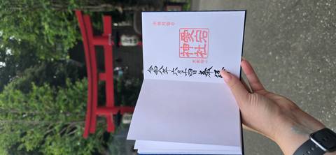       Hand holding shrine stamp book with red torii gate and stone steps blurred in background
  