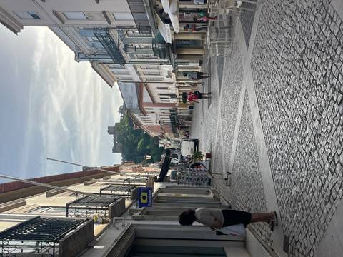       Wide cobbled street with distant hilltop castle, early morning light and locals walking
  