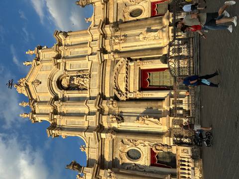       Baroque façade of Catania Cathedral warmly lit in evening light with pedestrians passing by
  