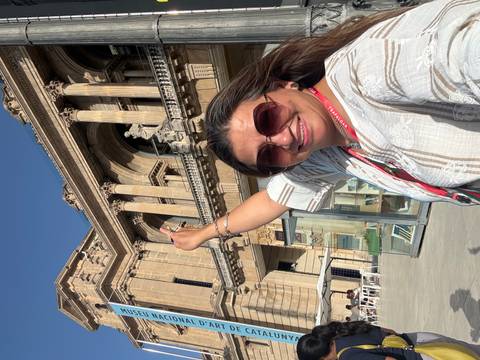       Woman taking a cheerful selfie with arm raised before a grand neoclassical facade in bright sunlight.
  