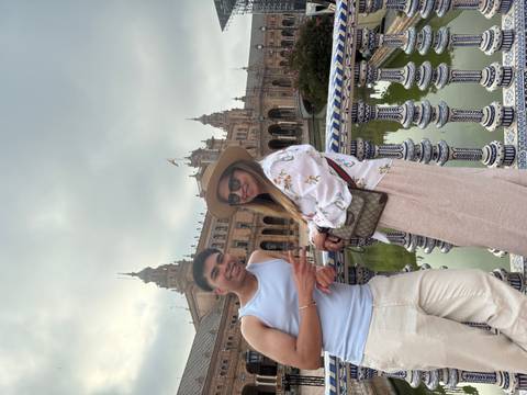       Two friends leaning against a tiled railing with Plaza de España’s grand architecture reflected in the canal behind.
  