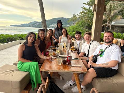       A smiling group enjoys sunset drinks at a beachside restaurant with palm trees and sea views.
  