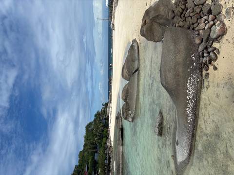       Clear shallows and smooth granite boulders line a quiet tropical beach under a partly cloudy sky.
  