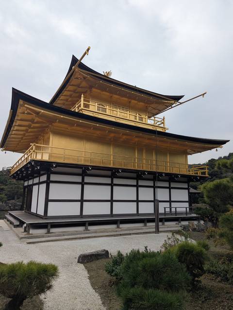       Side view of the gold-leaf-covered upper stories of Kinkaku-ji temple against a pale sky.
  
