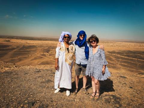       Two tourists and a local guide in traditional attire stand arm-in-arm on a desert ridge with dunes behind.
  