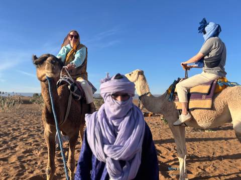       Travelers wrapped in scarves ride camels across sandy desert terrain with clear blue sky.
  