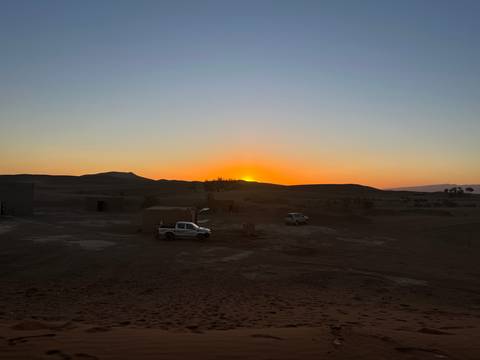       Pickup trucks parked on desert ground as the sun sets behind rolling dunes.
  