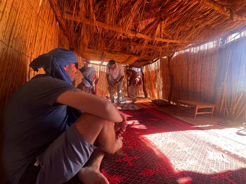       Travelers relax inside a thatched desert hut, sunlight filtering through woven walls.
  