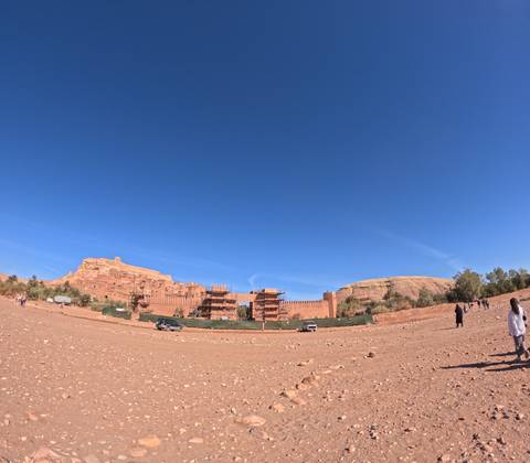       Panoramic shot of fortified clay buildings under scaffold near Ait Benhaddou with tourists scattered in foreground.
  