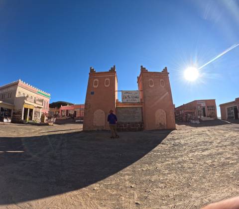       Traveler stands before twin adobe gate towers marking a desert town entrance under bright sun.
  