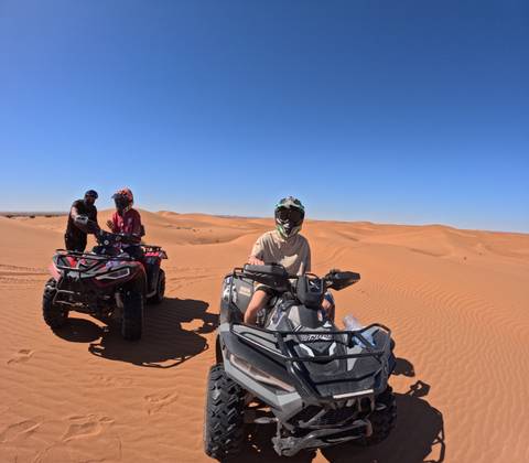       Adventurers on quad bikes pause atop rolling orange dunes under cloudless desert sky.
  