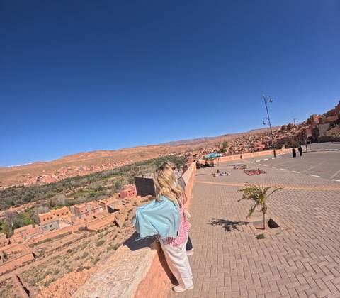       Woman with shawl looks out over terracotta town and arid hills from roadside terrace.
  