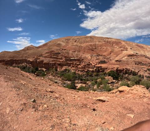      Reddish adobe ruins and scattered houses sit on a barren hillside above a green ravine in a dry mountain landscape.
  