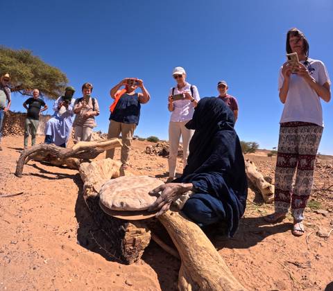       Travelers watch a veiled local woman bake flatbread over a stone in a sandy village clearing with a tree providing shade.
  