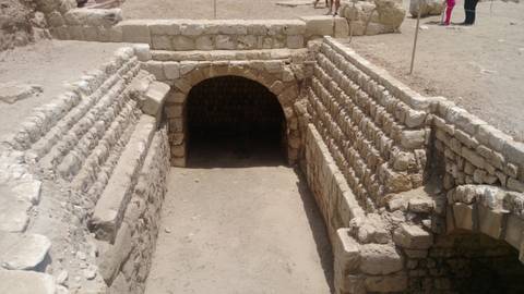       Stone-lined entrance to an ancient underground tomb or tunnel in the desert with visitors nearby.
  