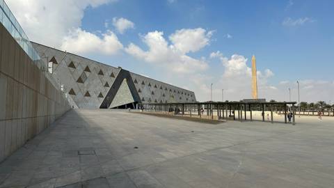       Expansive plaza of the Grand Egyptian Museum with triangular façade and obelisk under blue sky.
  