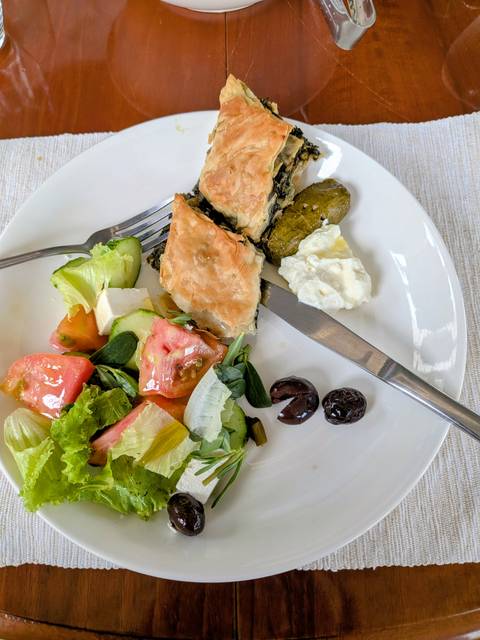       A plate features fresh salad, flaky pastry pie, olives and creamy cheese on a white tablecloth.
  