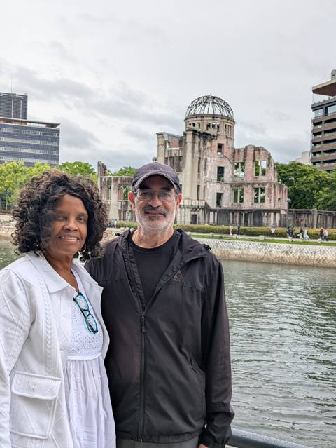       Two travelers stand beside a riverside view of the ruined Hiroshima Atomic Bomb Dome.
  