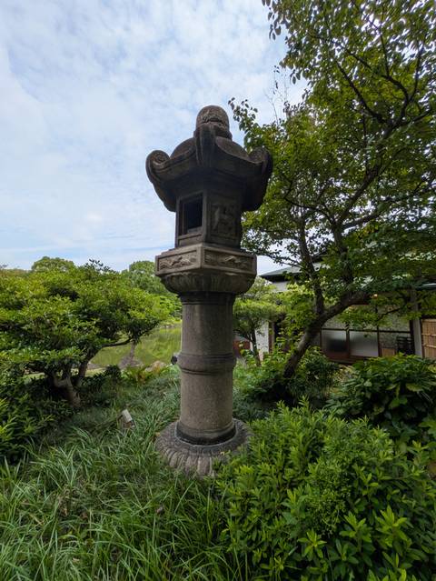       A stone lantern stands among manicured greenery beside a tranquil garden pond.
  