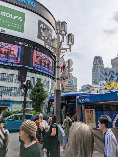      Digital billboards and modern towers dominate a busy Tokyo street scene near a bus stop.
  