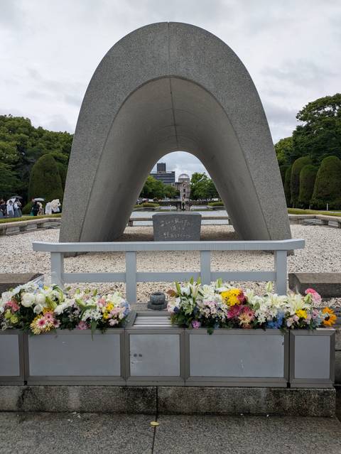       The arch cenotaph in Hiroshima Peace Park frames the Atomic Bomb Dome with fresh floral tributes in the foreground.
  
