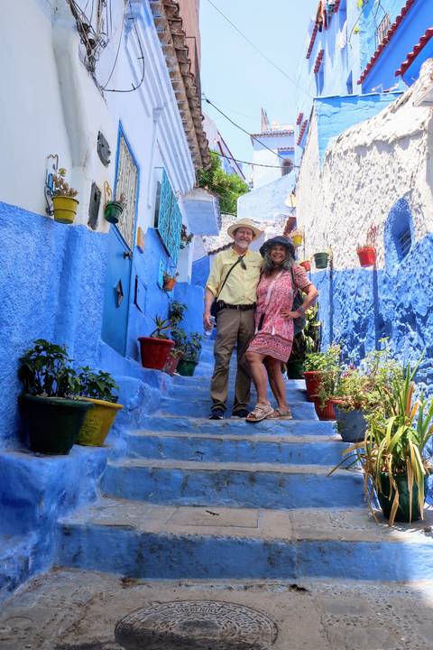       Smiling couple pose on blue-painted staircase surrounded by colourful potted plants in Chefchaouen alley
  