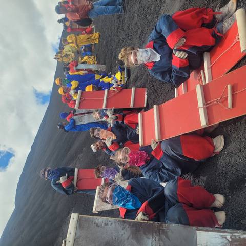       Adventurers in protective suits hold red boards on the dark ash slope of a volcano
  
