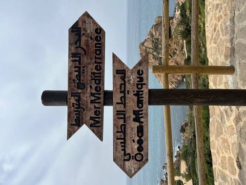      Bilingual wooden sign points toward the Mediterranean Sea and Atlantic Ocean with rocky coastline behind
  