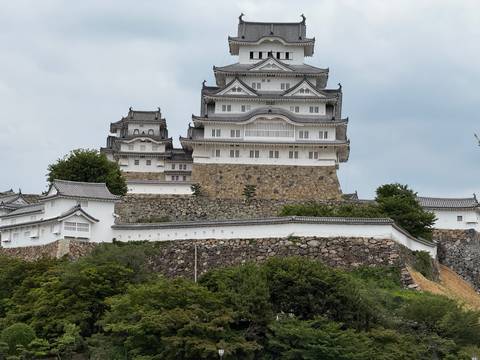       Majestic white-walled castle with layered roofs towers above stone ramparts and greenery.
  