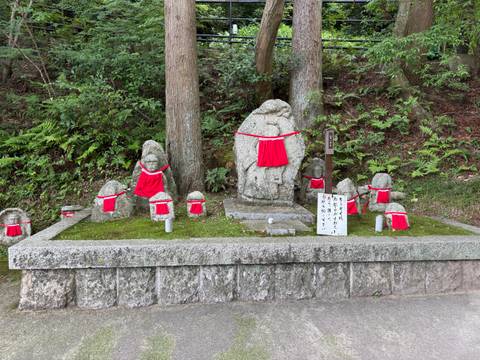       Row of small stone Jizo statues adorned with bright red bibs beneath tall trees.
  