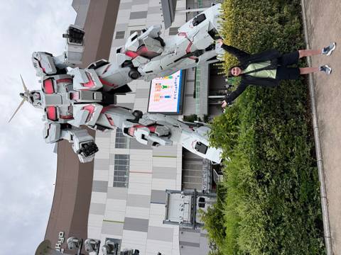       Traveller poses excitedly before a towering white Unicorn Gundam statue outside a mall.
  