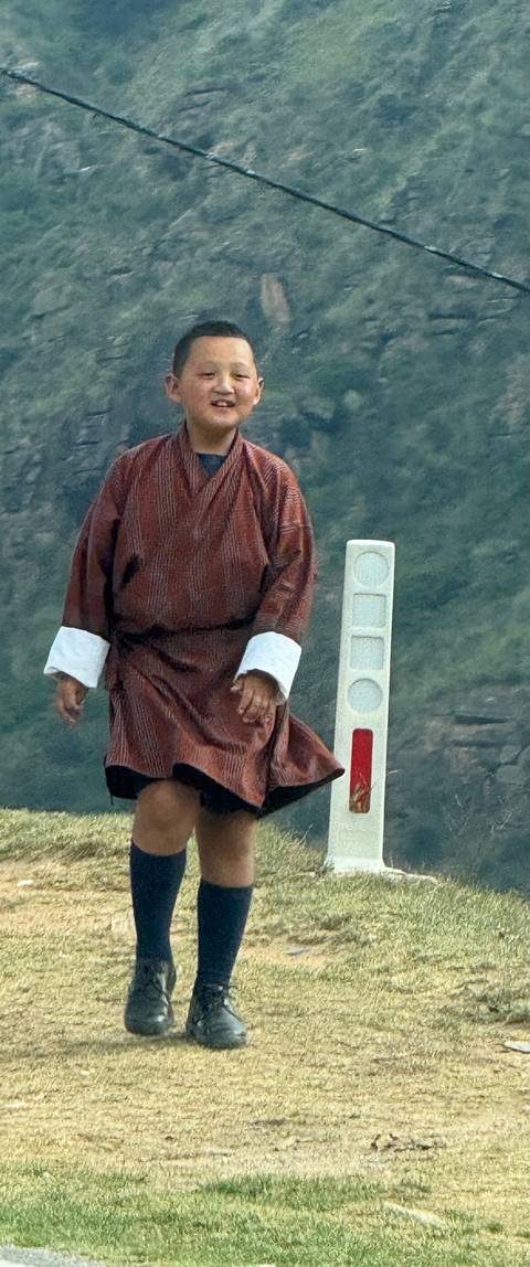      Bhutanese man in traditional gho walking beside a roadside marker against a misty mountain backdrop
  