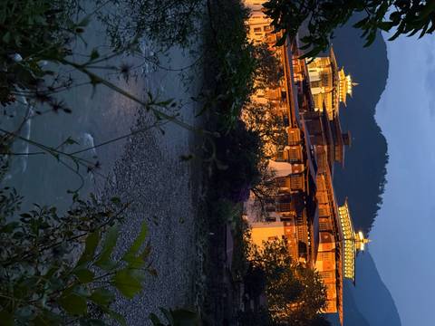       Illuminated Punakha Dzong monastery glowing at dusk beside a tranquil river with forested hills
  