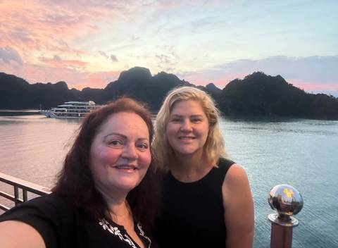       Two women taking a selfie on a boat in Halong Bay at sunset with limestone karsts behind.
  