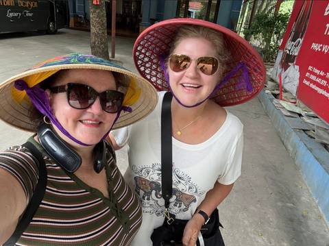       Two travelers wearing traditional conical hats and sunglasses smiling for a street selfie.
  