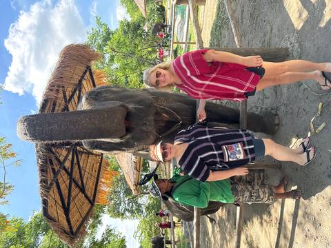       Two women posing beside a large elephant reaching its trunk toward the camera under a thatched shelter.
  