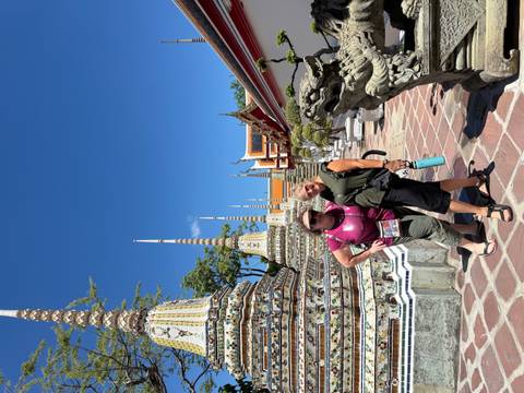       Two women posing in front of ornate, multi-coloured stupas inside a Thai temple complex under a bright blue sky.
  