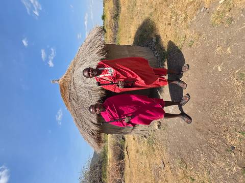       Two Maasai men in traditional red shukas stand outside a thatched hut.
  