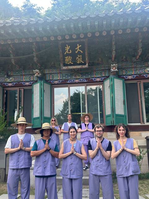       Group wearing traditional temple stay uniforms sit meditating in front of an ornately painted wooden building.
  