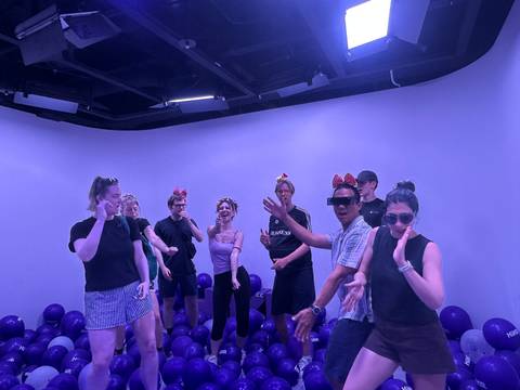       A group of young travelers dance and pose in a purple-lit room decorated with balloons and fun headbands.
  