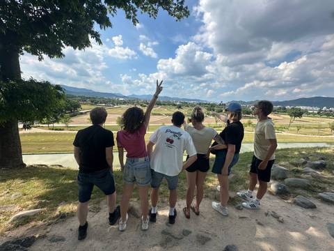       Six friends stand with their backs to the camera overlooking wide fields and distant hills under a bright blue sky.
  