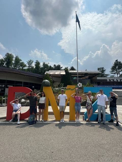       Travelers salute playfully in front of a colorful DMZ sign outside a visitor center on a bright day.
  