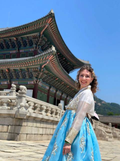       A traveler in a hanbok poses beside the ornate rooflines of Gyeongbokgung Palace under clear blue skies.
  