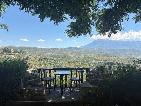       A shaded terrace with a table overlooks rolling valleys and distant mountains under a clear sky.
  