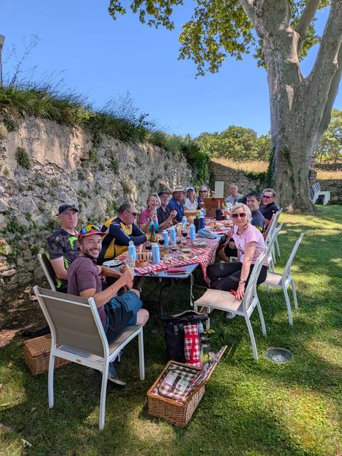      Large group enjoying an outdoor picnic lunch at a long table with checkered cloth beside a stone wall.
  