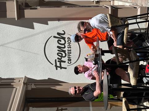       Cyclists relaxing with drinks at a sidewalk table outside a café that reads 'French Coffee Shop'.
  